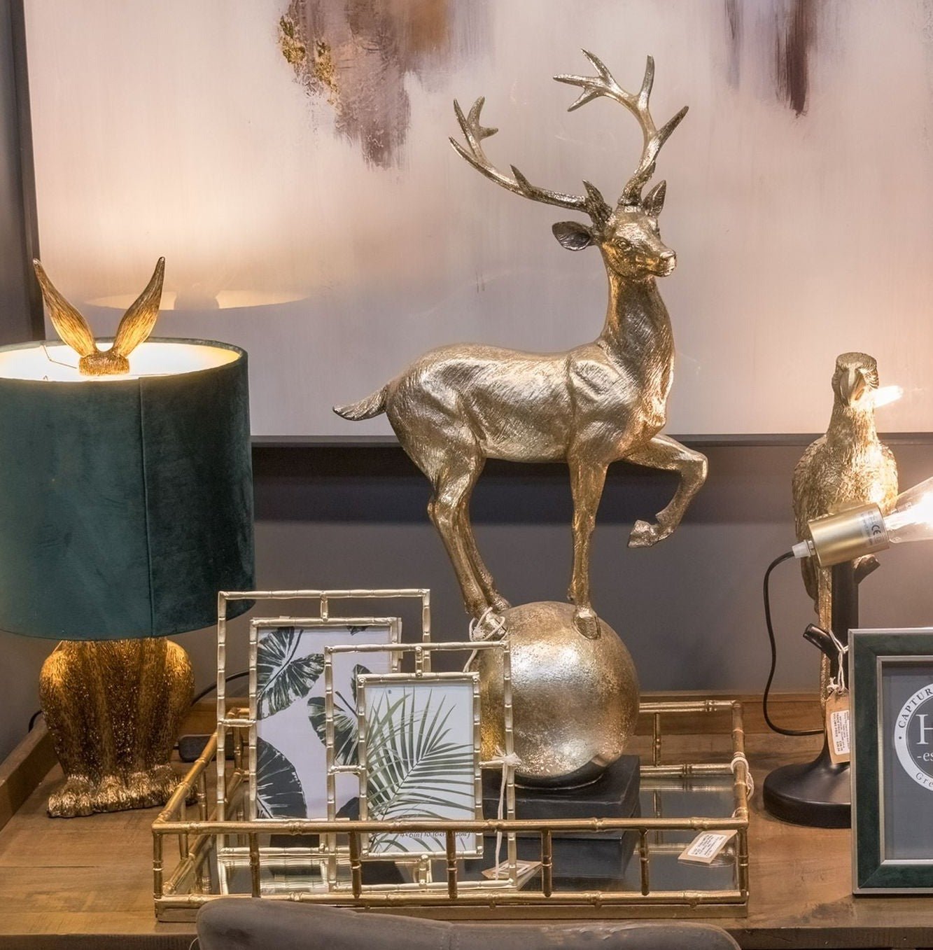 large rectangular tray on desk with two gold frames inside and stag ornament. A hare lamp with green shade is to the left and parrot lamp to the right