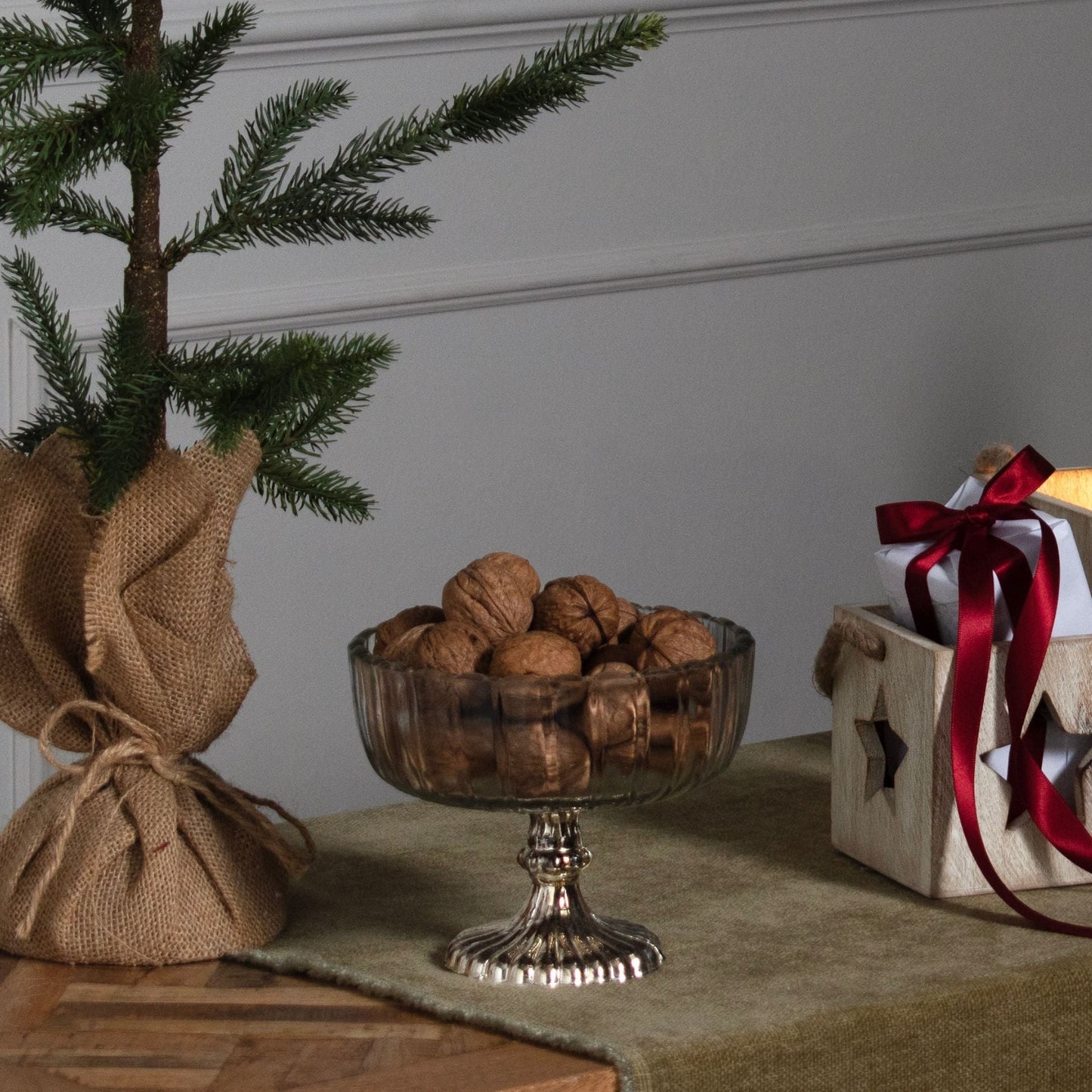 lifestyle image with walnuts in small fluted glass bowl. CHristmas tree in hessian sack to the left and small wooden box with star cut out to the right