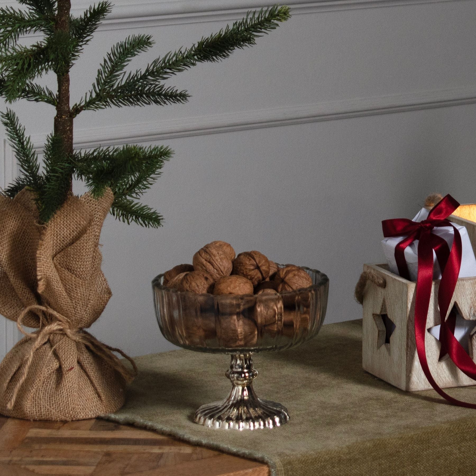 lifestyle image with walnuts in small fluted glass bowl. CHristmas tree in hessian sack to the left and small wooden box with star cut out to the right