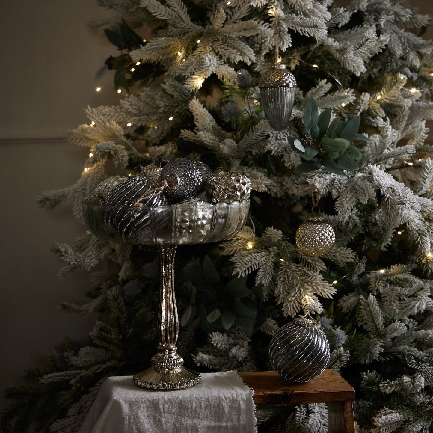 large fluted glass bowl with varying silver baubles in, in front of white Christmas tree decorated with silver baubles