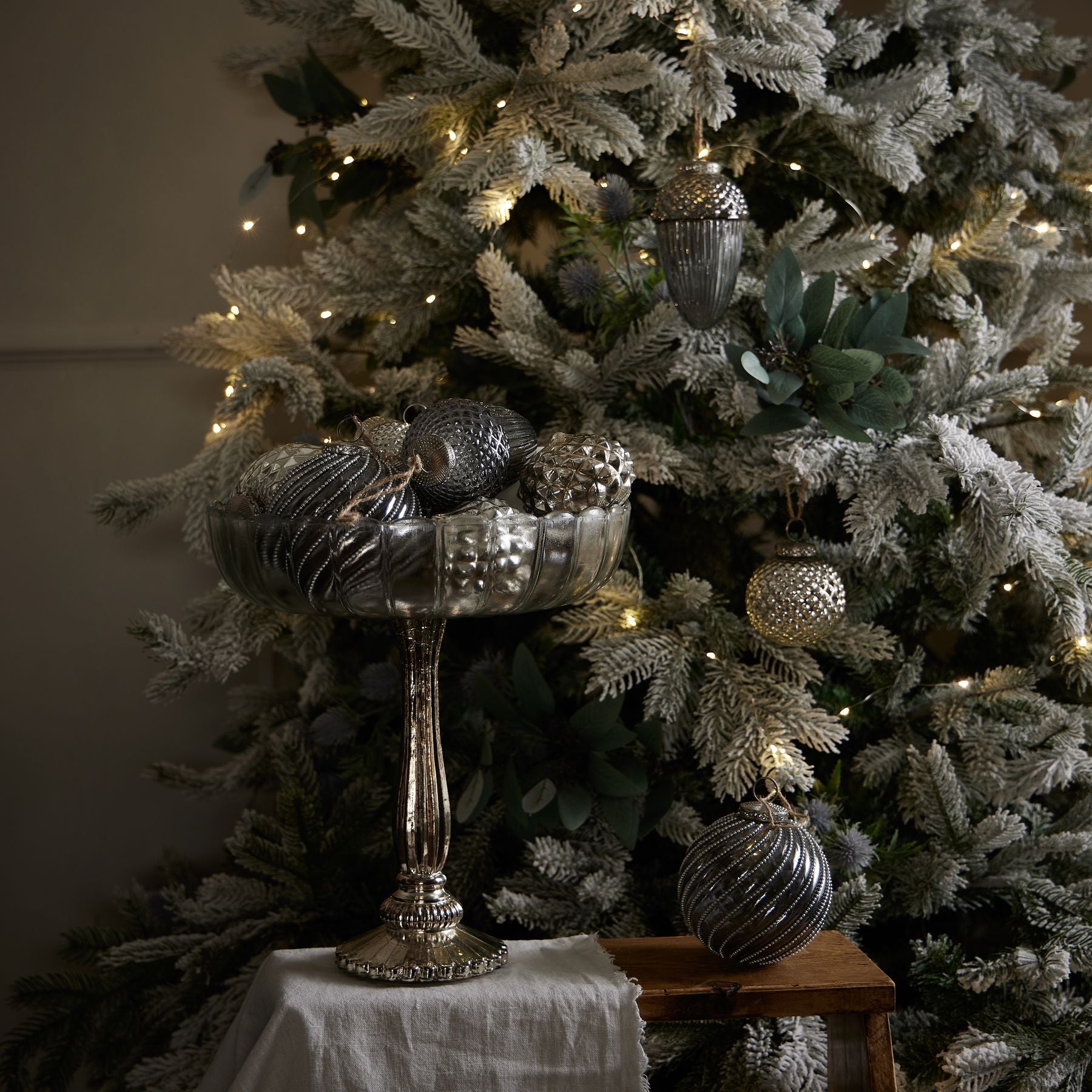 large fluted glass bowl with varying silver baubles in, in front of white Christmas tree decorated with silver baubles