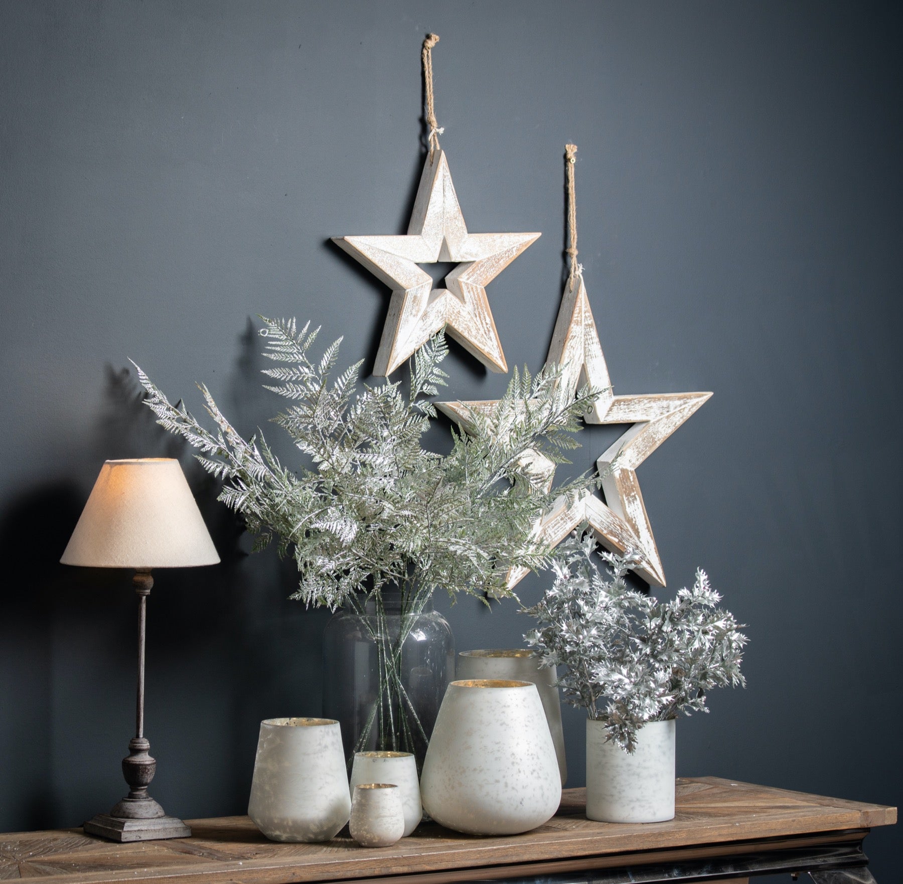 lifestyle image with both small and large wooden sparkle stars handing on dark grey wall. Underneath on a table sits a variety of white vases and Christmas themed greenery. A sleek wooden lamps sits to the left side