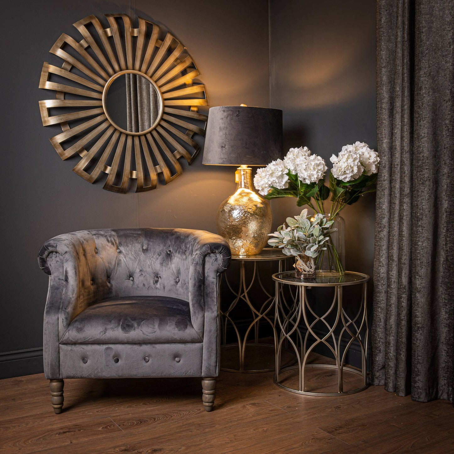 sunburst mirror in room set with chair, two tables, lamp on the larger table and artificial flowers on the smaller table in a clear glass vase
