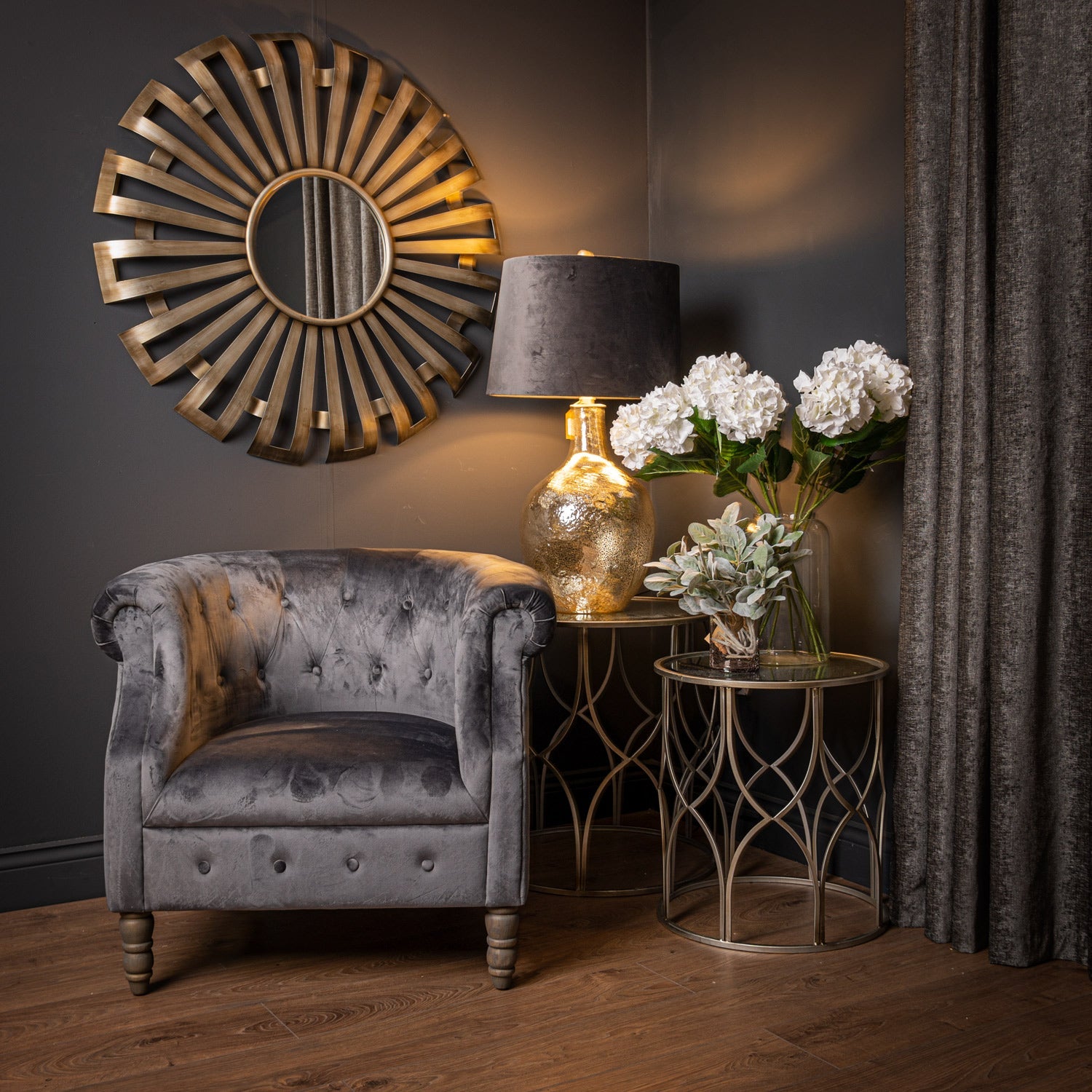 sunburst mirror in room set with chair, two tables, lamp on the larger table and artificial flowers on the smaller table in a clear glass vase