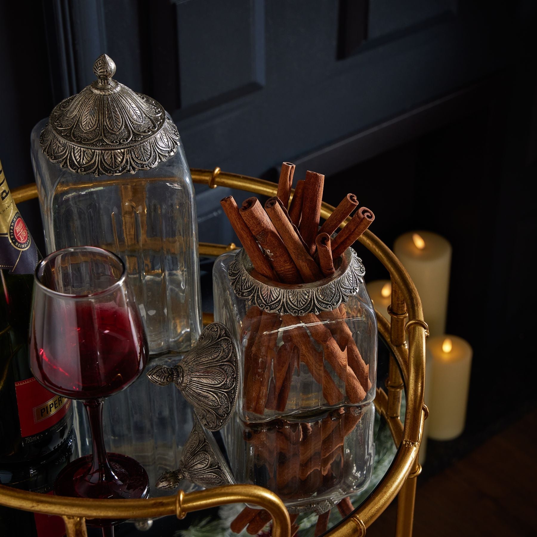 lifestyle image both small and large handblown glass jars on gold drinks trolley. The small jar has the lid off and contains cinnamon sticks, next to a glass of red wine. The large jar sits slightly behind with the lid on and is empty